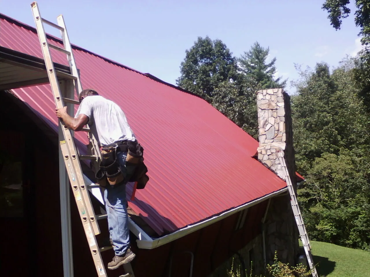 Roofer on a ladder beside a red metal roof and stone chimney — Soffit & Fascia Repair by Roofing Brothers in Prole